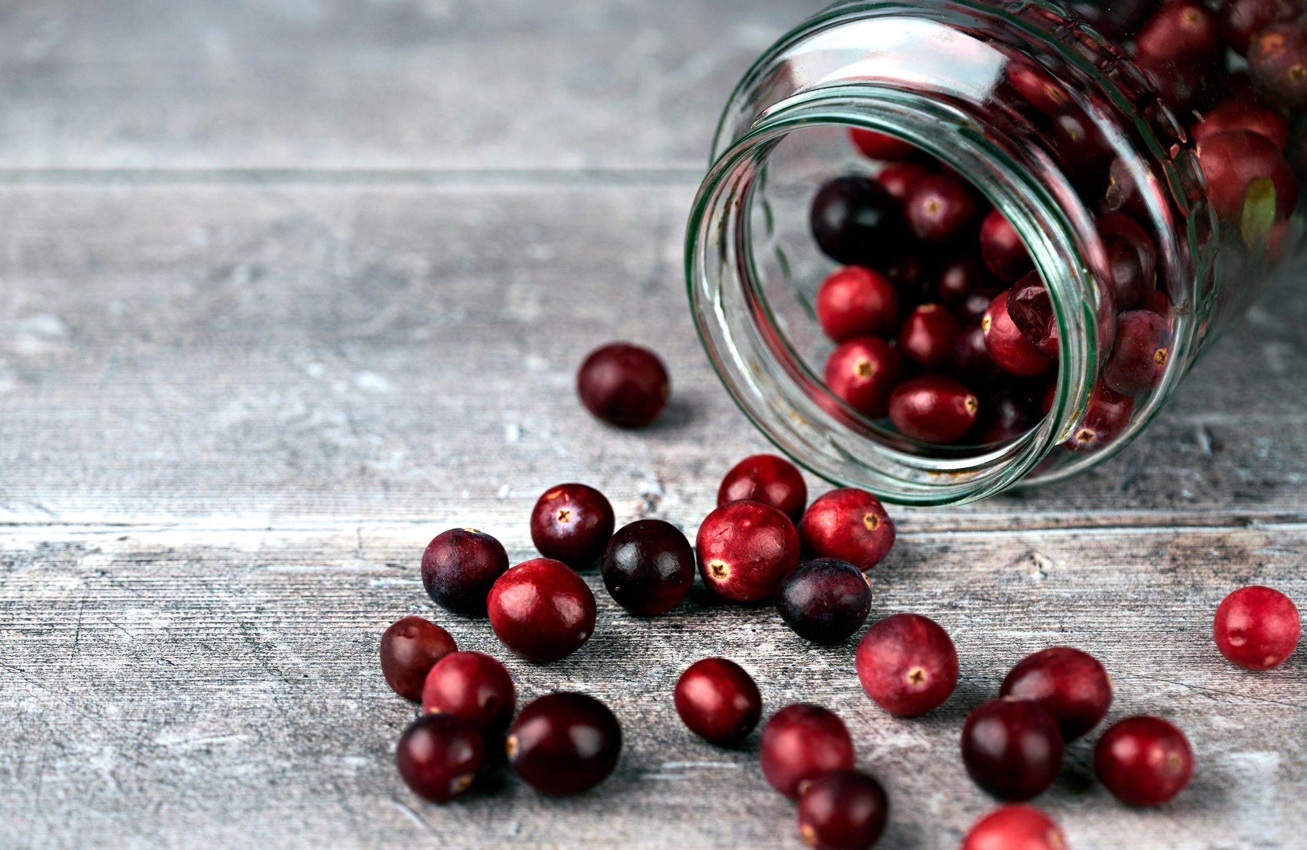 Fresh cranberries spilling out of a glass jar onto a rustic grey wooden surface, ready to be turned into homemade cranberry sauce.