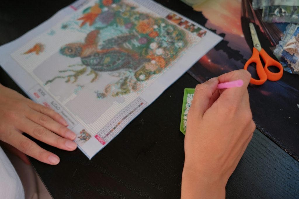 Hands placing diamond drills on a turtle diamond painting canvas using a pink applicator pen, with a bead tray and orange scissors on the table.