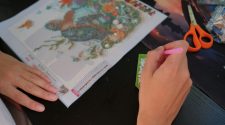 Hands placing diamond drills on a turtle diamond painting canvas using a pink applicator pen, with a bead tray and orange scissors on the table.