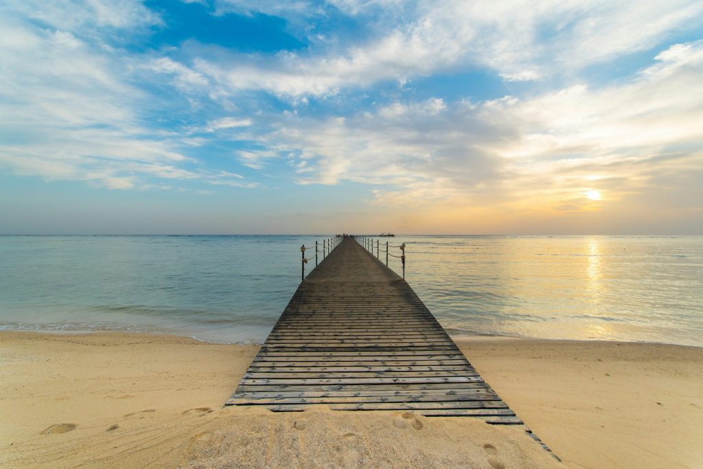 Wooden jetty stretching from a sandy beach into calm sea at sunrise, with soft clouds and warm golden light on the horizon.