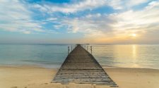 Wooden jetty stretching from a sandy beach into calm sea at sunrise, with soft clouds and warm golden light on the horizon.