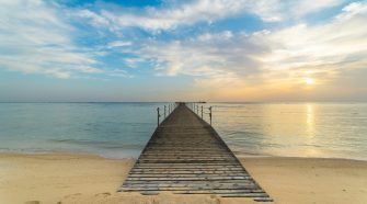 Wooden jetty stretching from a sandy beach into calm sea at sunrise, with soft clouds and warm golden light on the horizon.