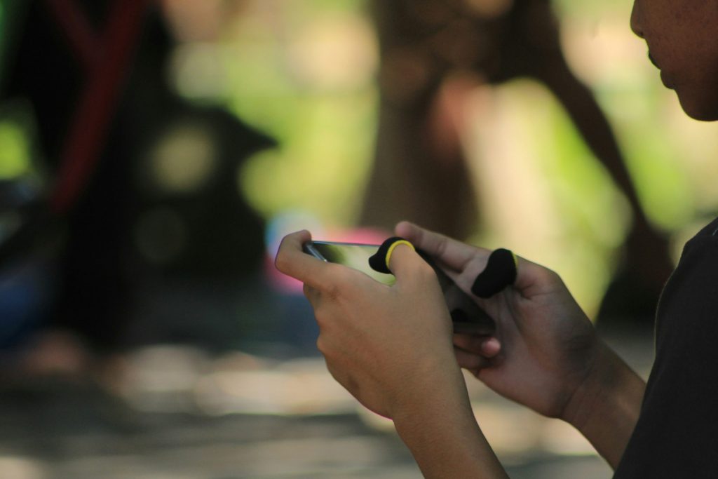 Close-up of someone holding a smartphone and gaming with finger sleeves, with a blurred outdoor background.