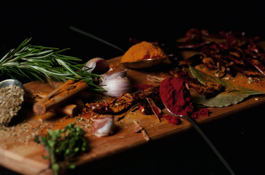 Rustic wooden board with taco seasoning ingredients including garlic, bay leaves, cinnamon, oregano, rosemary, turmeric, and paprika, styled against a dark background.