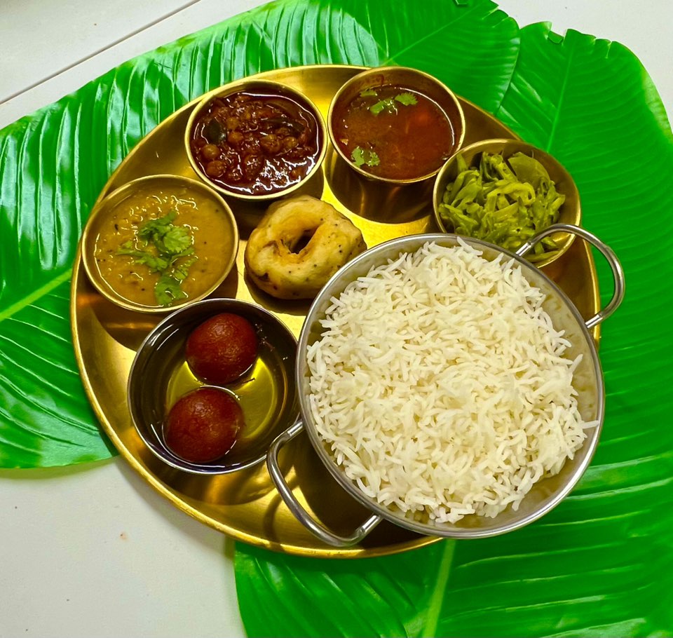 Traditional South Indian thali served on a brass plate with compartments holding white basmati rice, gulab jamun in syrup, dal, vegetable curry, sambar, chutney, medu vada, and a side of stir-fried green beans, placed on decorative green banana leaf placemats.