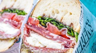 Alt text: Close-up of a freshly made Italian-style sandwich from Derek’s, filled with cured meats, mozzarella, tomato, lettuce, and pesto on crusty white bread.