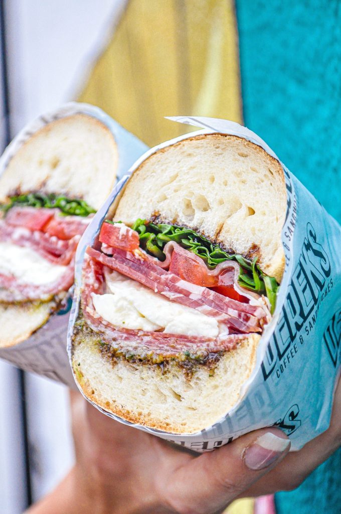 Alt text: Close-up of a freshly made Italian-style sandwich from Derek’s, filled with cured meats, mozzarella, tomato, lettuce, and pesto on crusty white bread.