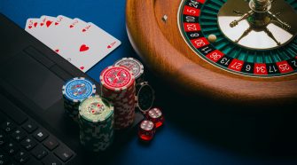 Poker chips and playing cards beside a laptop with a roulette wheel and dice on a blue table, representing online and casino gambling.
