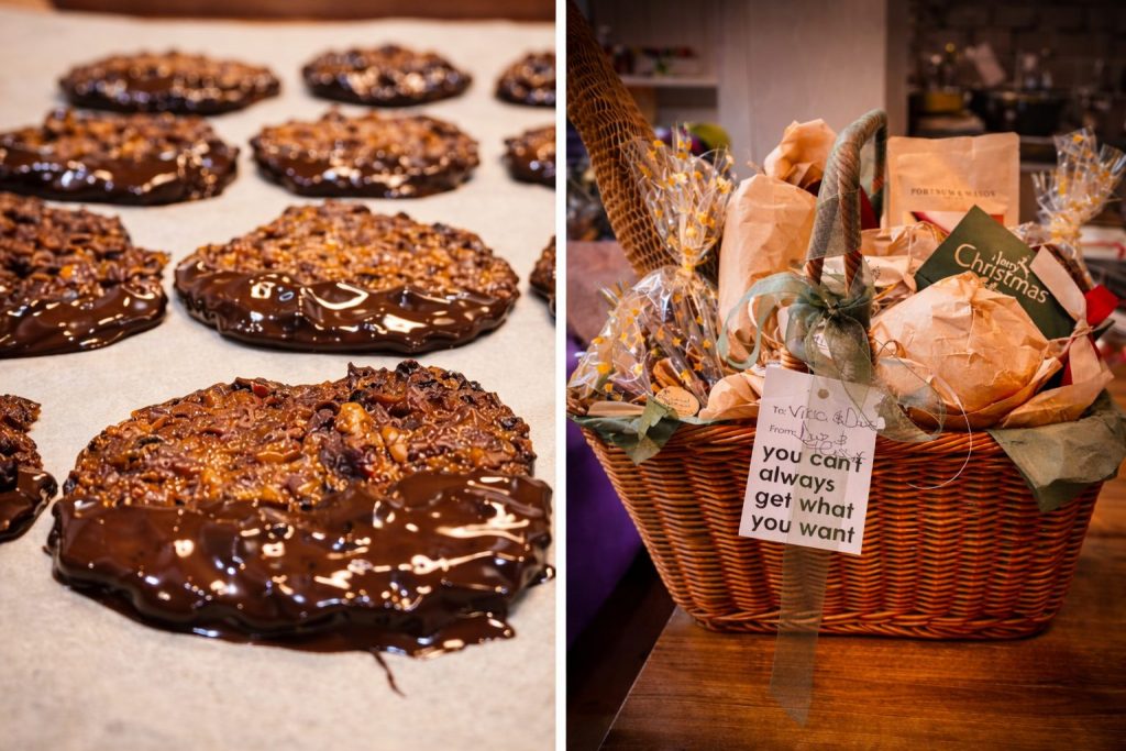 Close-up of glossy dark chocolate-dipped pecan florentines cooling on parchment, alongside a luxurious Fortnum & Mason-inspired Christmas hamper wrapped with ribbon, festive packaging, and handwritten gift tags.