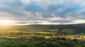 Rolling Yorkshire countryside at sunset, with patchwork green fields, stone walls, and moorland hills under a dramatic cloud-filled sky.