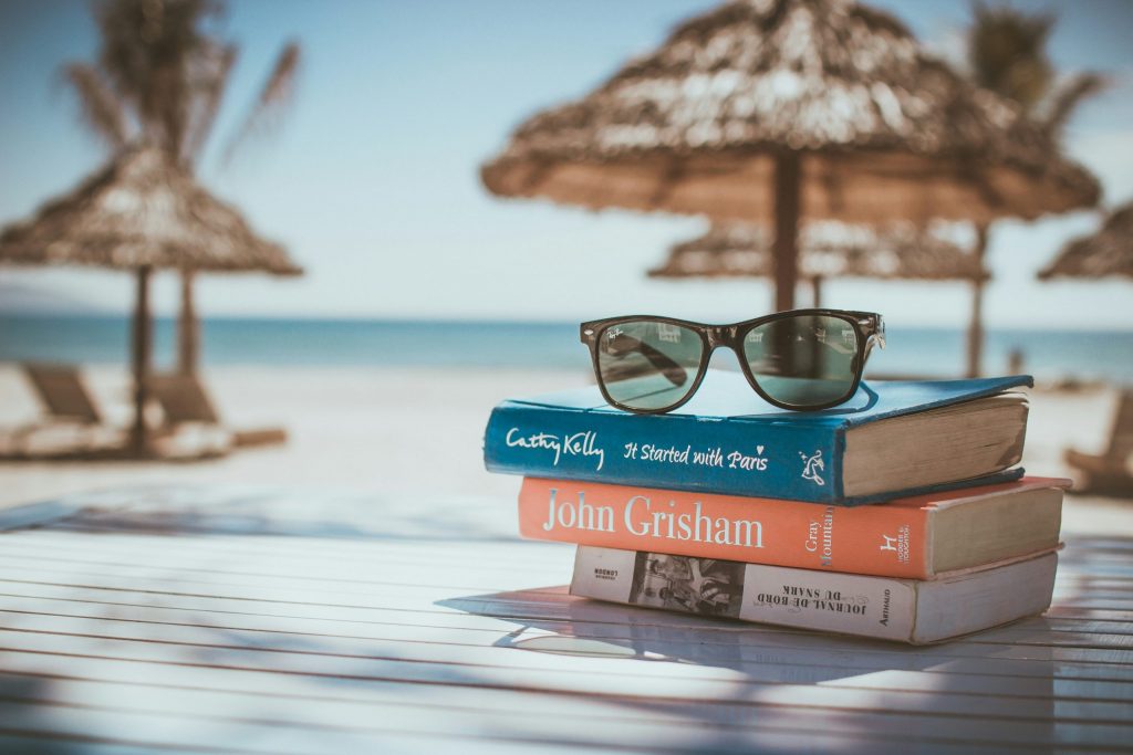 Stack of books with sunglasses on top sitting on a sunlit table by the beach, with parasols and the sea in the background, evoking a relaxed and affordable holiday atmosphere.