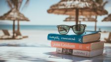 Stack of books with sunglasses on top sitting on a sunlit table by the beach, with parasols and the sea in the background, evoking a relaxed and affordable holiday atmosphere.