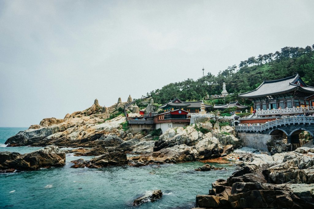Coastal Buddhist temple complex perched on rocky cliffs overlooking the sea in South Korea, with traditional Korean architecture, stone bridges, and turquoise water below.