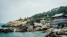 Coastal Buddhist temple complex perched on rocky cliffs overlooking the sea in South Korea, with traditional Korean architecture, stone bridges, and turquoise water below.