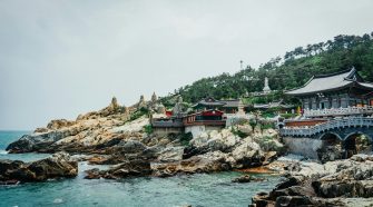 Coastal Buddhist temple complex perched on rocky cliffs overlooking the sea in South Korea, with traditional Korean architecture, stone bridges, and turquoise water below.
