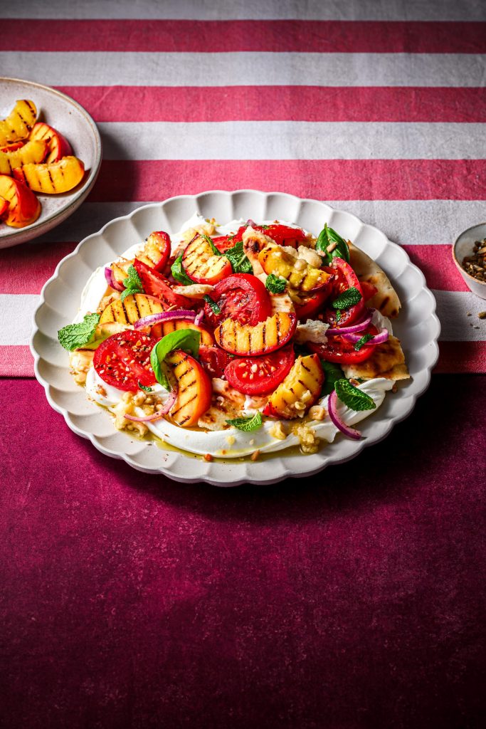 Grilled South African peach winter salad with tomatoes, labneh, fresh herbs and crisp flatbread served on a white platter.
