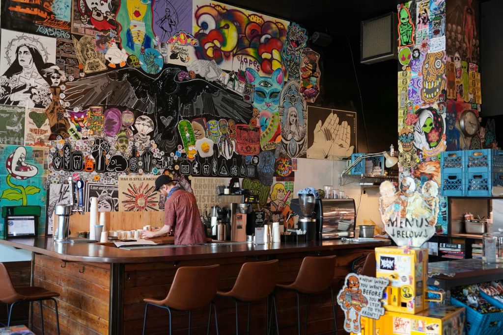 Alt text: Independent café interior with colourful street art walls, bar seating, and a staff member preparing drinks behind the counter, reflecting a modern, creative local dining space.