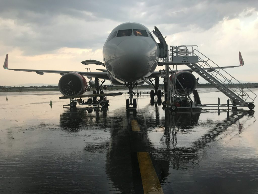 A commercial airplane on a wet airport runway during rainy weather, with passenger stairs positioned at the front and rear doors, and engine maintenance equipment nearby.