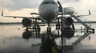 A commercial airplane on a wet airport runway during rainy weather, with passenger stairs positioned at the front and rear doors, and engine maintenance equipment nearby.