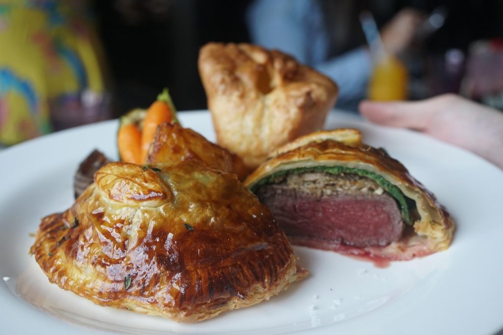 Individual beef Wellington parcels with golden puff pastry, one sliced open to reveal pink beef centre, served with roasted carrots and a Yorkshire pudding on a white plate.