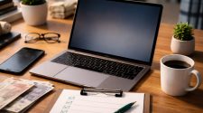 Laptop on a wooden desk with a smartphone, coffee mug, clipboard checklist, and pound notes, representing researching online casino safety and verification before gambling.