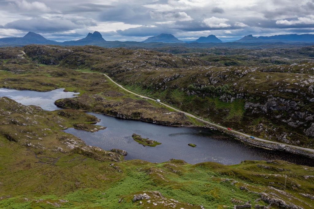 Aerial view of a winding road through the rugged Scottish Highlands along the North Coast 500, with lochs, rocky landscape and distant mountains under dramatic clouds.