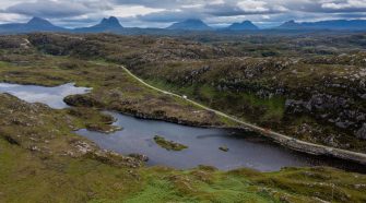 Aerial view of a winding road through the rugged Scottish Highlands along the North Coast 500, with lochs, rocky landscape and distant mountains under dramatic clouds.