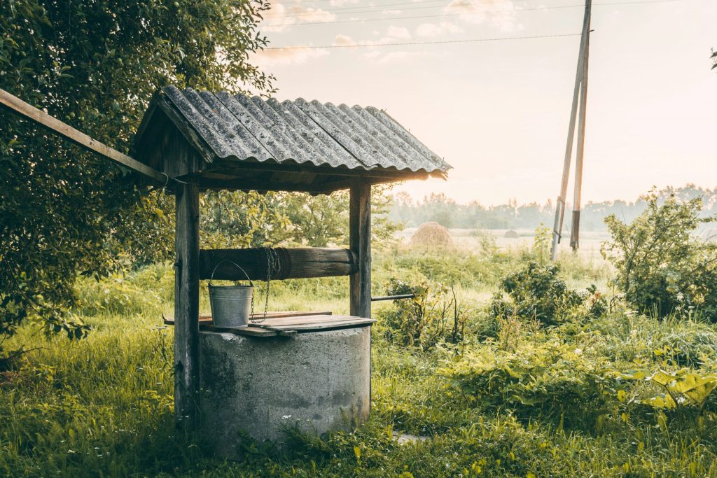Rustic covered water well with bucket in a grassy rural landscape at sunrise.