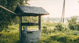 Rustic covered water well with bucket in a grassy rural landscape at sunrise.