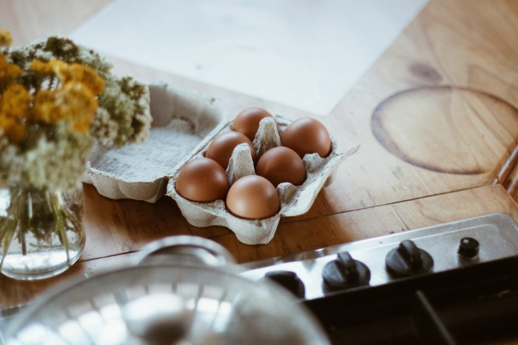 Carton of brown eggs on a wooden kitchen worktop beside a chopping board and stove, representing everyday grocery staples in a home setting.