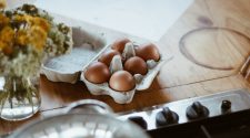 Carton of brown eggs on a wooden kitchen worktop beside a chopping board and stove, representing everyday grocery staples in a home setting.