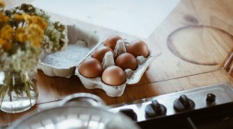Carton of brown eggs on a wooden kitchen worktop beside a chopping board and stove, representing everyday grocery staples in a home setting.
