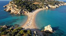 Aerial view of a curved sandy beach in Sicily with turquoise waters, rocky cliffs, and sunbathers along the shoreline.