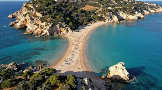 Aerial view of a curved sandy beach in Sicily with turquoise waters, rocky cliffs, and sunbathers along the shoreline.