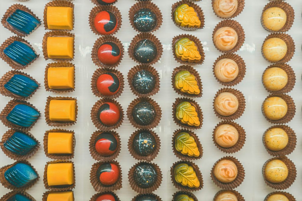Overhead view of assorted artisan chocolates arranged in neat rows, featuring colourful glossy bonbons in blue, yellow, red, and speckled designs, each placed in individual paper cases.