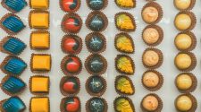 Overhead view of assorted artisan chocolates arranged in neat rows, featuring colourful glossy bonbons in blue, yellow, red, and speckled designs, each placed in individual paper cases.