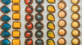 Overhead view of assorted artisan chocolates arranged in neat rows, featuring colourful glossy bonbons in blue, yellow, red, and speckled designs, each placed in individual paper cases.