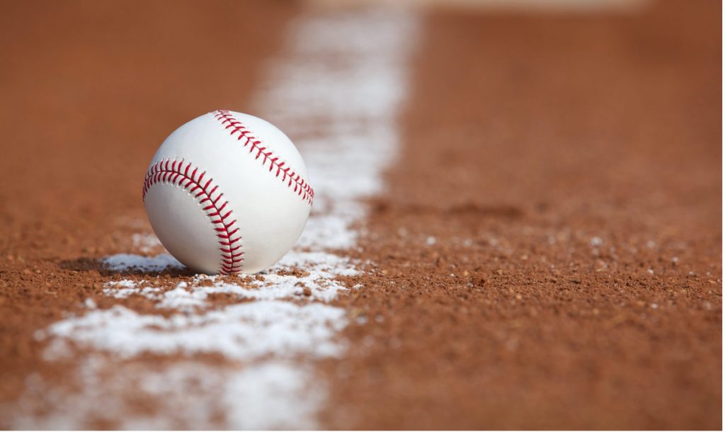 Close-up of a white baseball resting on the brown infield dirt beside a chalk line, with the background softly blurred.