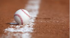 Close-up of a white baseball resting on the brown infield dirt beside a chalk line, with the background softly blurred.