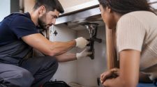 A plumber kneels beneath a modern kitchen sink using a wrench to repair pipework while a woman watches closely beside him in a bright, contemporary home.