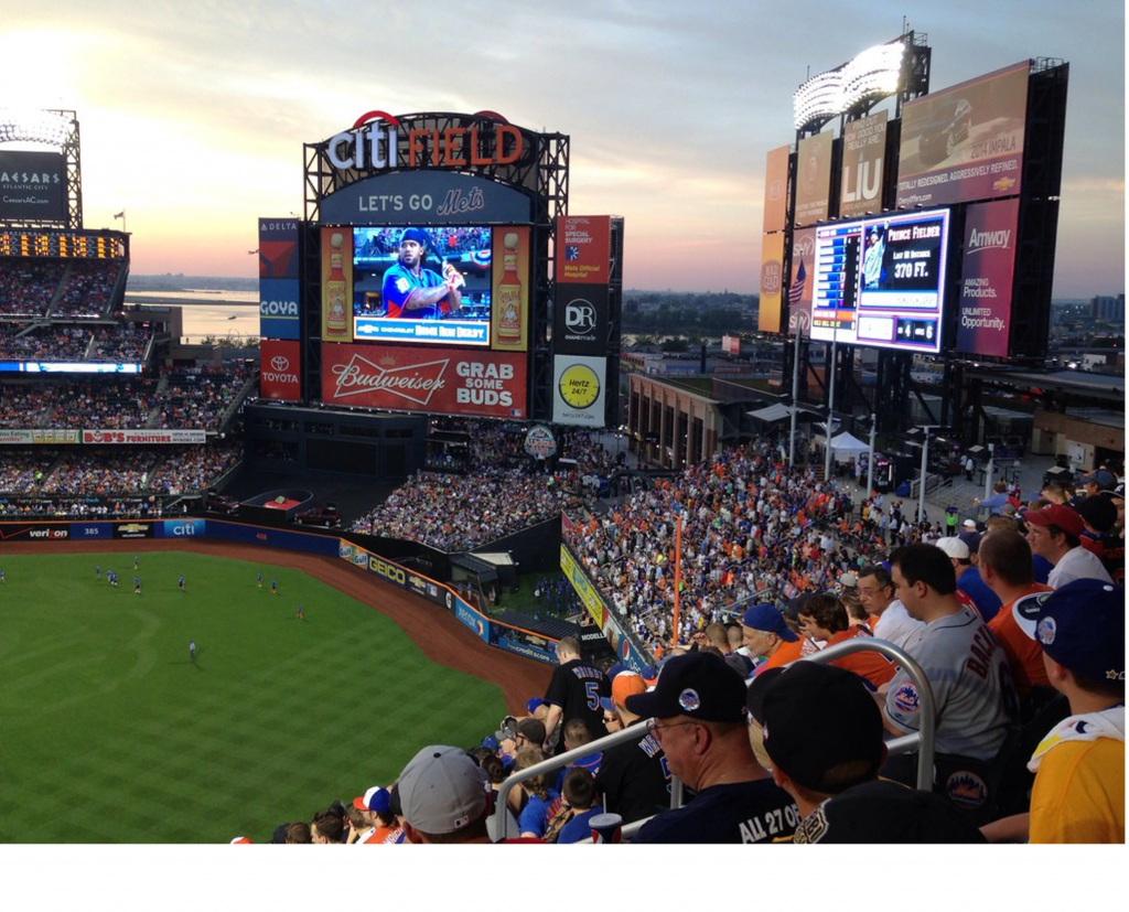 Wide view of Citi Field at sunset during a New York Mets baseball game, with a packed crowd, glowing scoreboard and the field below.