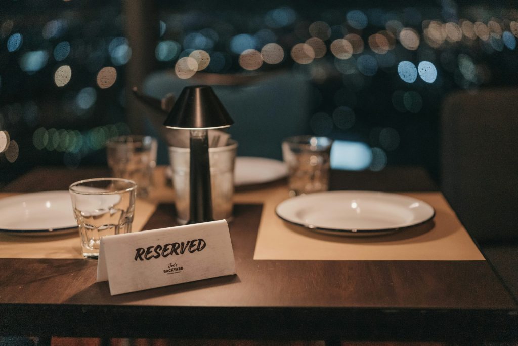Reserved table for two in a softly lit restaurant, with plates, glasses and a small lamp against a blurred city lights backdrop.