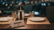 Reserved table for two in a softly lit restaurant, with plates, glasses and a small lamp against a blurred city lights backdrop.