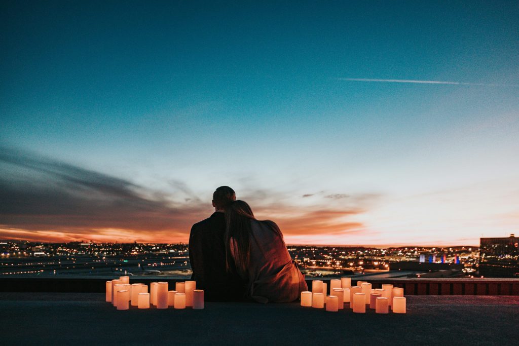 A couple sits together on a rooftop at sunset, surrounded by glowing candles and overlooking a city skyline.