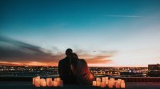A couple sits together on a rooftop at sunset, surrounded by glowing candles and overlooking a city skyline.
