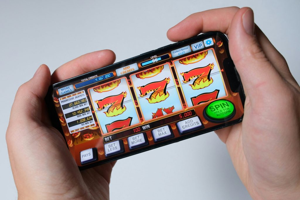 Hands holding a smartphone displaying an online slot machine game with flaming number sevens and a green spin button.