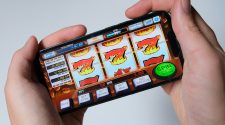 Hands holding a smartphone displaying an online slot machine game with flaming number sevens and a green spin button.