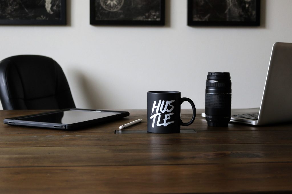 Desk setup with a laptop, tablet, camera lens, stylus, and a black “hustle” mug, representing freelance work, creativity, and side hustle productivity.
