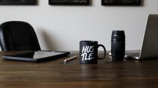 Desk setup with a laptop, tablet, camera lens, stylus, and a black “hustle” mug, representing freelance work, creativity, and side hustle productivity.