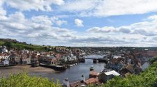 Whitby harbour viewed from above on a bright day, showing the River Esk, colourful rooftops, fishing boats, and the historic bridge under a dramatic cloud-filled Yorkshire sky.